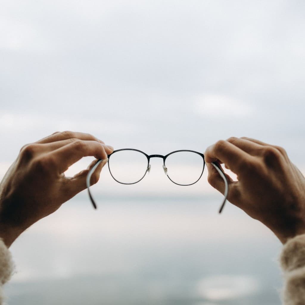 Person holding a pair of eyeglasses up to a soft, cloudy sky, symbolizing clarity of vision through clean and well-maintained lenses.