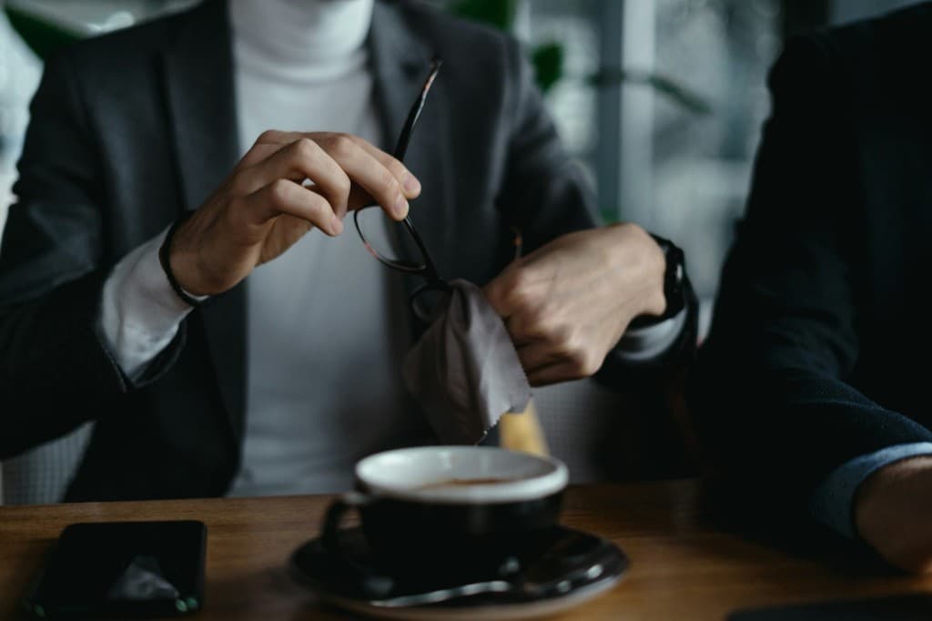 Person cleaning eyeglasses with a soft cloth at a table, showing the proper way to wipe lenses without causing scratches.