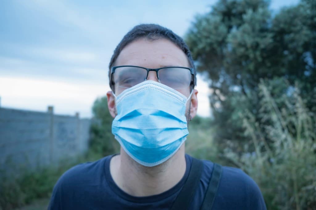 Person outdoors wearing a blue medical mask and glasses fogged from warm breath rising into the lenses due to limited airflow.