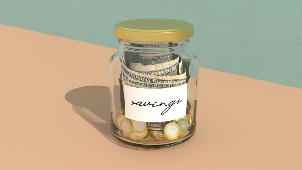 Glass jar labeled savings filled with cash and coins on a plain background, symbolizing saving money.